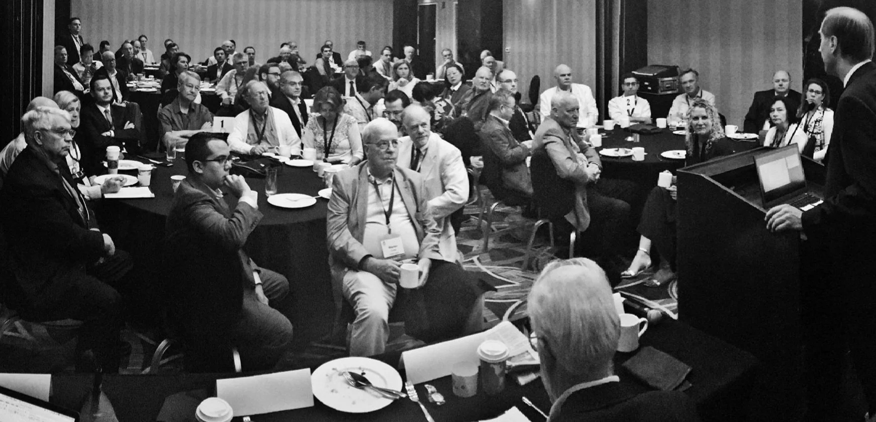 A conference audience seated at round tables, listening to a speaker at a lectern